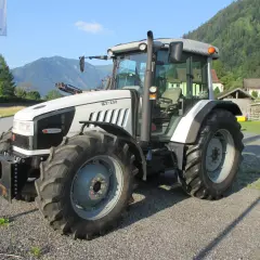 Biały traktor Hurlimann XT-130 stoi na żwirowej drodze, w tle góry i domy.