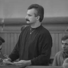 Mężczyzna z wąsami, niczym minstrel, trzyma dokumenty w sali sądowej. Obok niego siedzą ludzie w mundurach.