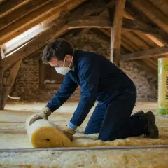 Un ouvrier installe un isolant jaune dans un grenier. Le rouleau d'isolant et la mention "prix isolation toiture" sur le produit suggèrent une rénovation pour améliorer le confort thermique.