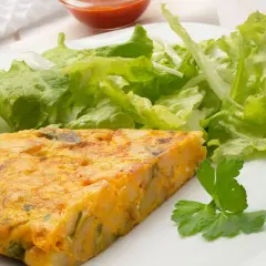 Una porción de tortilla de patatas, dorada y jugosa, acompañada de una fresca ensalada verde y un toque de perejil.