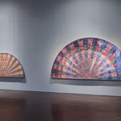 Two fan-shaped artworks display intricate pattern and decoration, creating a sense of movement. The larger piece bursts with colorful fabric squares, while the smaller one features a softer, sunburst design.