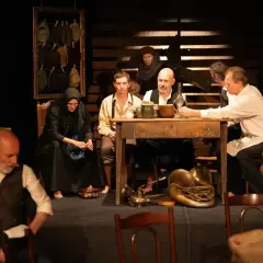 Scena teatralna, grupa ludzi przy stole, atmosfera napięcia i oczekiwania. Ten dramat porusza trudne tematy.