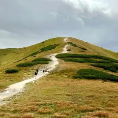 Wędrówka po górskim szlaku, jedna z wielu atrakcji w Zakopanem. Ludzie idą ścieżką na tle zielonych wzgórz i chmur.