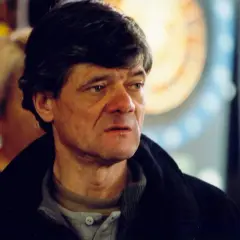 Mężczyzna o zmęczonej twarzy, w ciemnej kurtce, w tle rozmyte postacie i okrągły obiekt.