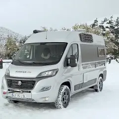 Camping-car 4x4 argenté garé dans la neige, avec des arbres enneigés en arrière-plan.