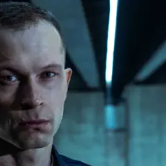 Zbliżenie na twarz mężczyzny z filmu "Tomasz Komenda". Jego spojrzenie jest intensywne, a tło sugeruje mroczne otoczenie.