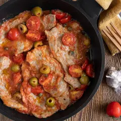Thinly sliced meat in a tomato sauce with olives and cherry tomatoes in a pan.