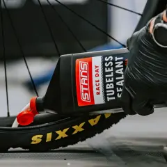 Gloved hands pour Stans Race Day tubeless sealant into a Maxxis tire, demonstrating how to add sealant to tubeless tires for a puncture-free ride.