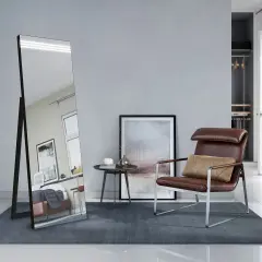 Eleganckie, duże lustro stojące w nowoczesnym wnętrzu, odbijające łóżko i okno. Obok fotel i stolik.