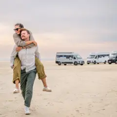 Szczęśliwa para na plaży, mężczyzna niesie kobietę na barana. W tle kilka kamperów, czyli vanów mieszkalnych, gotowych na przygodę.