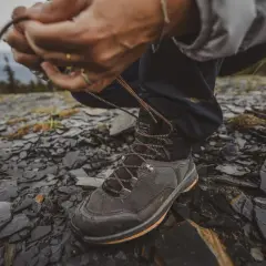 Ręce zawiązują sznurówki wysokich butów trekkingowych na kamienistym, wilgotnym podłożu.