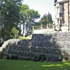 Imponująca skarpa na granicy działki, zbudowana z kamieni w gabionach, tworzy tarasy porośnięte zielenią. Na górze widać fragment rezydencji.