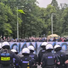 Policja w pełnym rynsztunku stoi przed grupą pseudokibiców, którzy trzymają transparent.