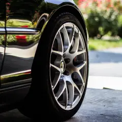 Detalle de un coche negro con llantas plateadas, listo para cambiar ruedas delanteras. Reflejos de flores y vegetación en la carrocería.