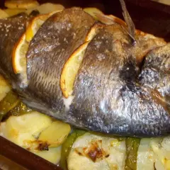 Dorada al horno con rodajas de limón, patatas y pimientos verdes en una bandeja.
