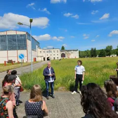 Gruppe bei einem **Festival im Osten** vor Industriegebäuden. Ein Mann erklärt etwas, während die Besucher zuhören.