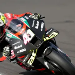 Piloto de MotoGP inclinado en una curva, con su casco blanco y rosa y la moto negra con detalles amarillos.