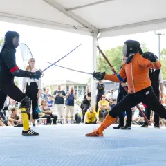 Två personer i skyddsutrustning utövar hema kampsport med svärd. Publiken tittar på.