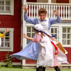 Barn leker vid Junibacken Madicken hus. En vuxen i blå klänning och två barn i tidsenliga kläder.