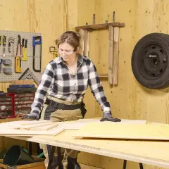En kvinna arbetar med plywood utomhus i en verkstad. Verktyg hänger på väggen, och däck är upphängda.