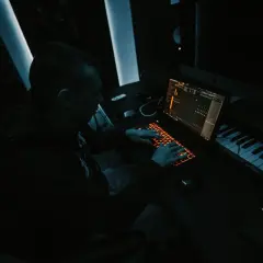 Producent muzyczny tworzy przebój w ciemnym studiu, pracując przy laptopie i klawiaturze sterującej.