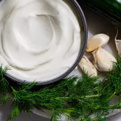 Crema blanca cremosa en un tazón, rodeada de eneldo fresco, ajos y un pepino cortado. Ingredientes para una deliciosa salsa tzatziki.