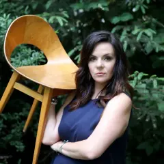 Krystyna Łuczak-Surówka, w wieku, z dumą prezentuje designerskie krzesło na tle zieleni.