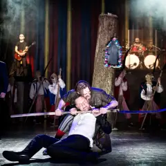 Jan Klata spektakle: dramatyczna scena z kosami i muzyką na żywo. Dwóch mężczyzn walczy na scenie, jeden klęczy, drugi go dusi.