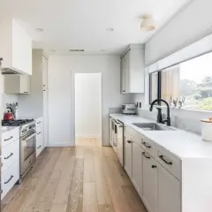 A bright, modern galley kitchen with white cabinets, stainless steel appliances, and a large window overlooking greenery.