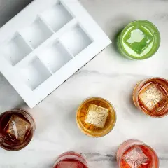 En vit form för klar is bredvid glas med drinkar. Varje drink har en stor, klar isbit med ett mönster.