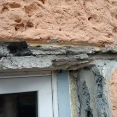 Uszkodzony montaż nadproża w starym domu, widoczne zbrojenie i luźne elementy betonowe nad oknem.
