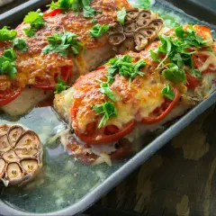 Aromatyczny dorsz pieczony w piekarniku z pomidorami i serem, ozdobiony świeżą natką pietruszki i pieczoną główką czosnku.