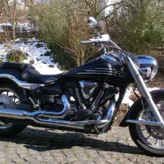 Błyszczący czarny motocykl Yamaha Raider 1900, z chromowanymi detalami, zaparkowany na bruku. W tle zima i krzewy.