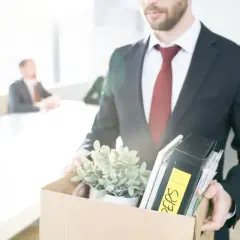 Un homme en costume quitte son bureau, portant une boîte remplie d'objets personnels et de dossiers. Il vient de déposer sa lettre de démission période d'essai.