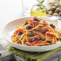 Makaron spaghetti z pomidorami, oliwkami i kaparami, podany w białej misce na zielonej serwetce.