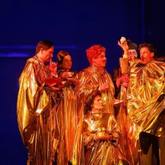 Aktorzy w złotych szatach, trzymający filiżanki i książki, tworzą scenę z najlepszych spektakli teatralnych.