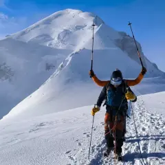 Un alpiniste victorieux lève ses bâtons sur fond de Mont Blanc. L'ascension est réussie, la neige brille sous le ciel bleu.
