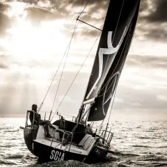 Vela nera con logo, un uomo a bordo di una barca a vela che naviga in mare con un cielo drammatico. L'imbarcazione mostra un angolo di deriva accentuato.