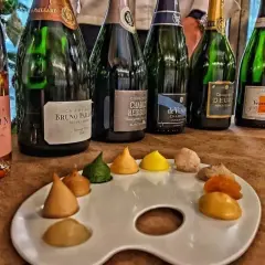 Bottiglie di champagne, tra cui Veuve Clicquot e Bruno Paillard, disposte su un tavolo con una tavolozza da pittore piena di dolcetti colorati, un'ode al gusto e all'arte del cote de bar.