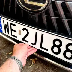 Dłoń poprawia tablicę rejestracyjną auta. Wartość samochodu powyżej 150 tys. zł, widoczna na grillu i zderzaku.