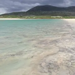 Vackert turkost vatten möter en sandstrand med mönster från tidvattnet. Hus och en grön kulle syns i bakgrunden på Yttre Hebriderna.