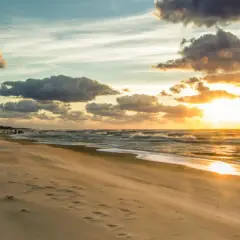 Zachód słońca nad Bałtykiem, plaża i falochron. W oddali widać ludzi spacerujących i kitesurferów. To idealne miejsce na podziwianie atrakcji Jastrzębiej Góry i okolic.