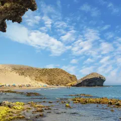 Vista de la playa de San José con rocas cubiertas de algas en primer plano y gente disfrutando del sol bajo sombrillas.
