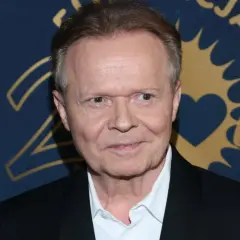 Michał Bajor w czarnym garniturze i białej koszuli, z partnerem u boku, pozuje na tle złoto-granatowej ścianki.