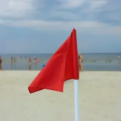 Czerwona flaga nad morzem oznacza zakaz kąpieli. Na plaży widać ludzi, ale woda jest niebezpieczna.