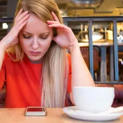 Zmartwiona kobieta z rękami na skroniach, zastanawia się, czy kawa podnosi ciśnienie. Obok telefon i filiżanka.