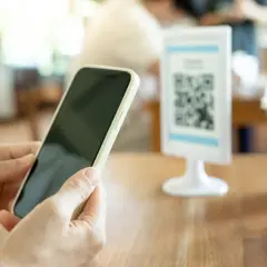 Ręce trzymają telefon z Androidem, gotowe do zeskanowania kodu QR na stoliku w kawiarni.
