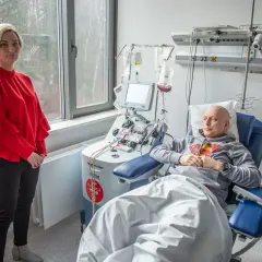 Kobieta w czerwonej bluzce stoi obok pacjenta podłączonego do aparatury medycznej. To ważny moment w procesie przeszczepu szpiku.