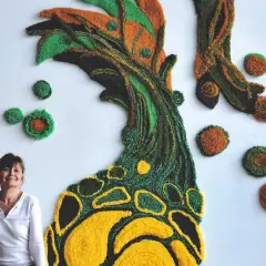 Une designer textile pose devant sa création murale en relief, un arbre aux couleurs vives.