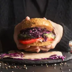 Ręce trzymają soczystego burgera z czerwoną kapustą, pomidorem i ogórkiem. Idealny przykład, jak składać burgera.